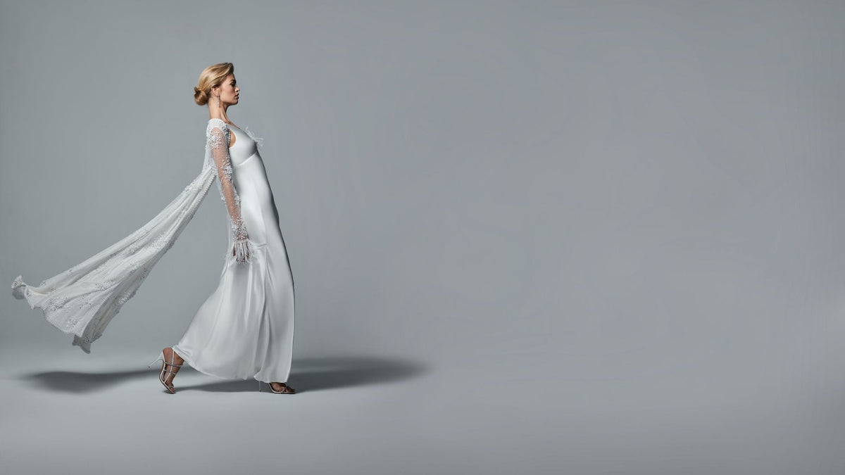 Side profile of a model wearing a long white gown with sheer embellished sleeves and a flowing cape train in a studio setting